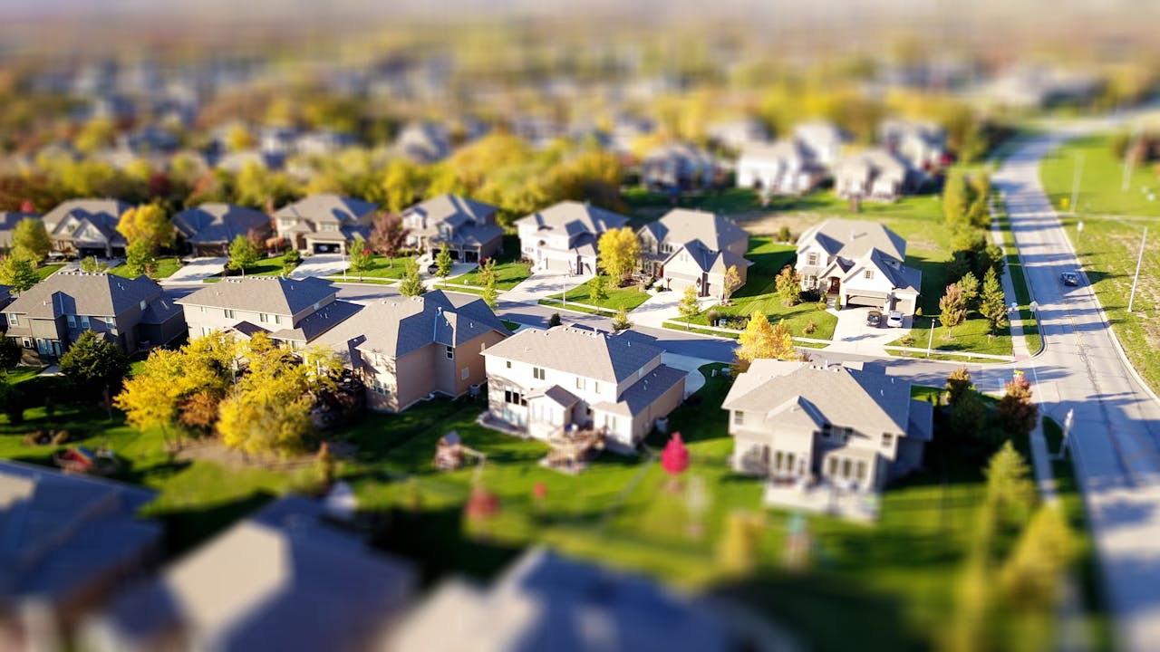 story-img Tilt-shift aerial photo of a suburban neighborhood with green lawns and roads.